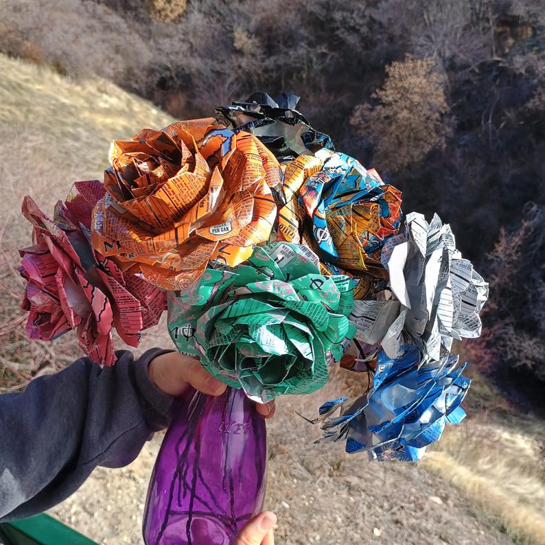May include: A bouquet of handmade paper roses in various colors, including orange, pink, green, blue, and silver. The flowers are crafted from recycled materials and arranged in a purple glass vase. The background shows a natural outdoor setting.