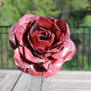 May include: A large, handmade rose made from recycled red and white printed material. The flower is in full bloom, with detailed petals and a clear glass vase. The background is a blurred outdoor scene with green foliage.