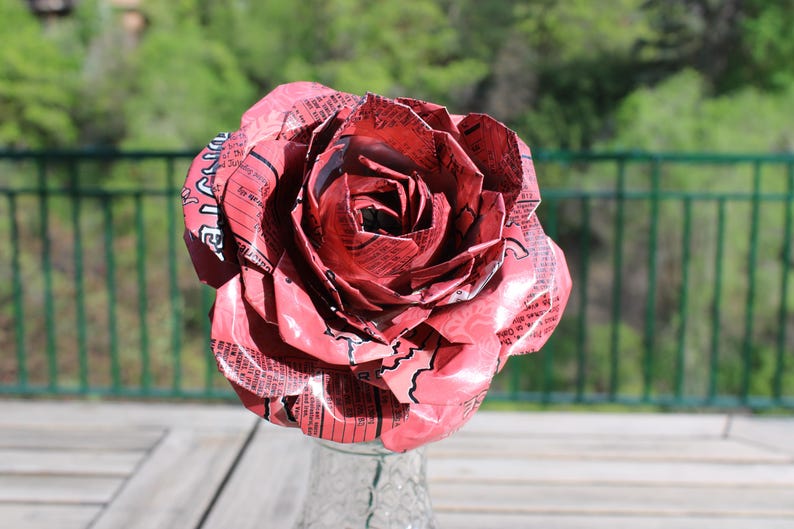 May include: A large, handmade rose made from recycled red and white printed material. The flower is in full bloom, with detailed petals and a clear glass vase. The background is a blurred outdoor scene with green foliage.