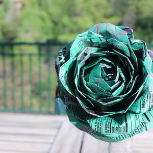 May include: A handmade paper rose in shades of green and teal, crafted from recycled paper. The flower's petals are intricately folded, showcasing the texture and detail of the repurposed material. The background is a blurred view of a natural outdoor setting.