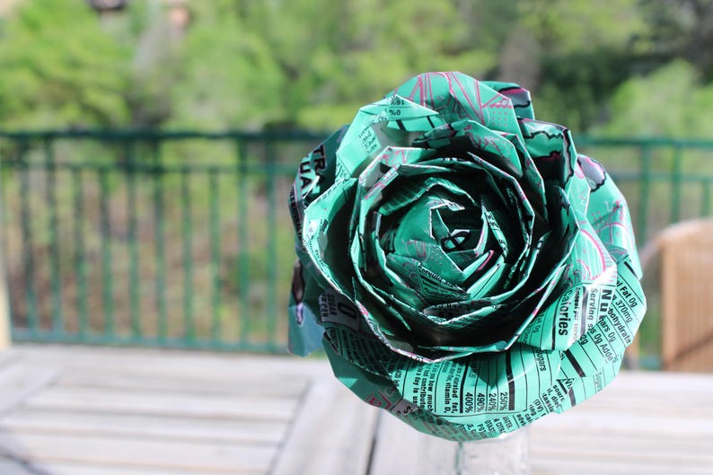 May include: A handmade paper rose in shades of green and teal, crafted from recycled paper. The flower's petals are intricately folded, showcasing the texture and detail of the repurposed material. The background is a blurred view of a natural outdoor setting.