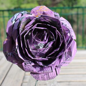 May include: A large, handmade purple rose made from recycled materials. The flower's petals are intricately folded, revealing text and patterns from the original packaging. The rose is a unique, eco-friendly decorative item.