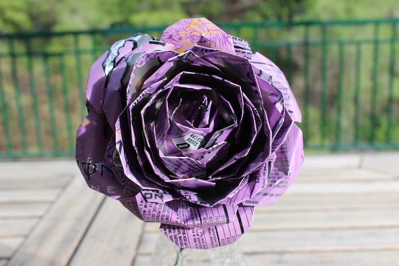 May include: A large, handmade purple rose made from recycled materials. The flower's petals are intricately folded, revealing text and patterns from the original packaging. The rose is a unique, eco-friendly decorative item.