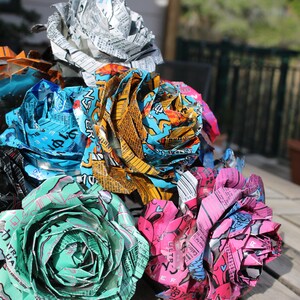 May include: A collection of handmade paper roses crafted from colorful aluminum cans. The flowers display a variety of colors, including orange, blue, green, pink, and silver. The roses are arranged on a wooden surface.