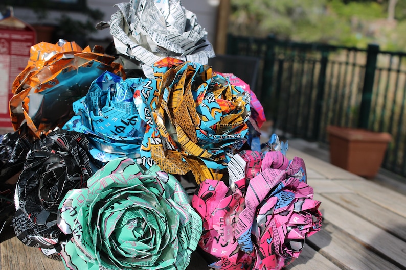 May include: A collection of handmade paper roses crafted from colorful aluminum cans. The flowers display a variety of colors, including orange, blue, green, pink, and silver. The roses are arranged on a wooden surface.