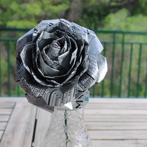 May include: A handmade paper rose made from recycled materials, displayed in a clear glass vase. The flower's petals are crafted from printed paper, creating a unique, eco-friendly decorative item. The vase is on a wooden table.