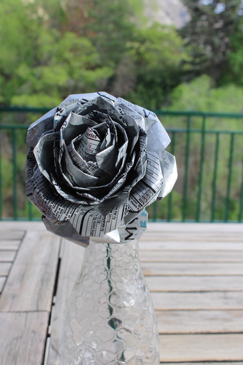 May include: A handmade paper rose made from recycled materials, displayed in a clear glass vase. The flower's petals are crafted from printed paper, creating a unique, eco-friendly decorative item. The vase is on a wooden table.