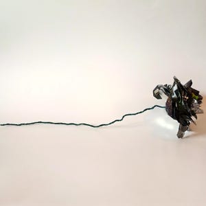 May include: A handmade paper flower with a dark green stem. The flower head is made of folded, multi-colored paper, with shades of green, black, and silver. The stem is a thin, dark green wire.