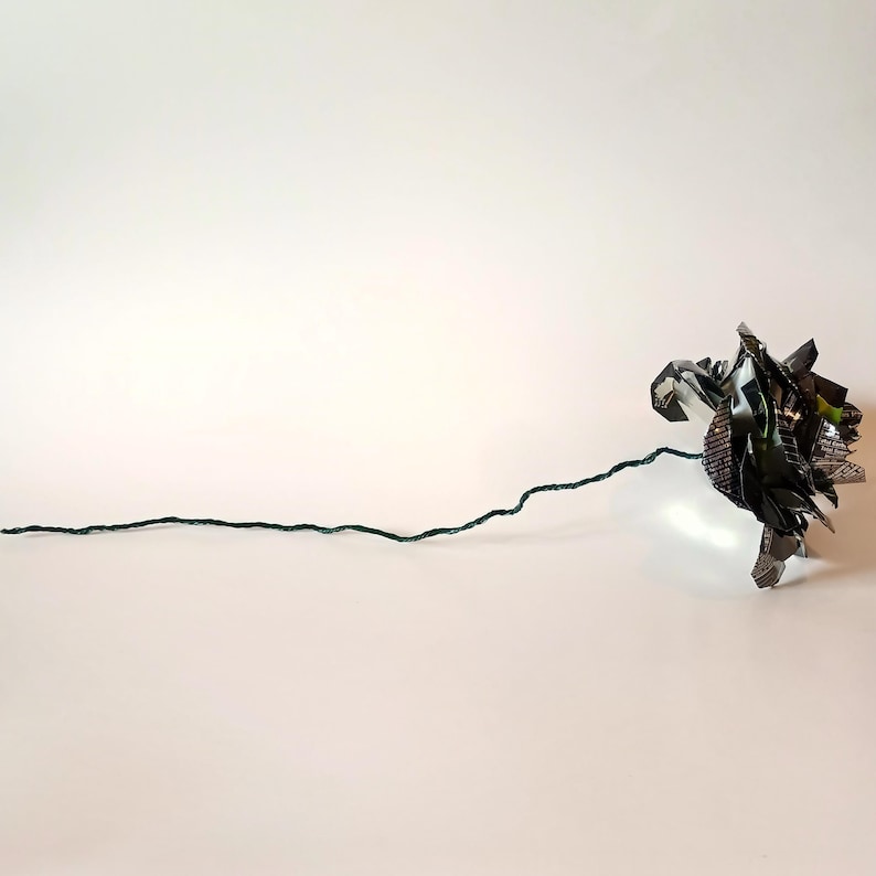 May include: A handmade paper flower with a dark green stem. The flower head is made of folded, multi-colored paper, with shades of green, black, and silver. The stem is a thin, dark green wire.