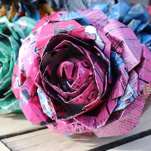 May include: Close-up of handmade paper roses in shades of pink, green, blue, and red. The petals are crafted from recycled materials, showcasing a unique texture and design. The roses are arranged on a wooden surface.