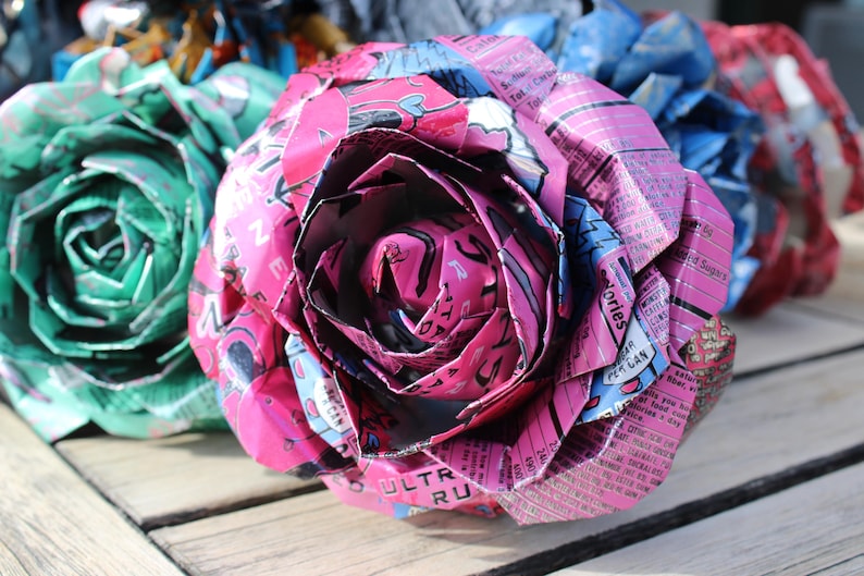May include: Close-up of handmade paper roses in shades of pink, green, blue, and red. The petals are crafted from recycled materials, showcasing a unique texture and design. The roses are arranged on a wooden surface.