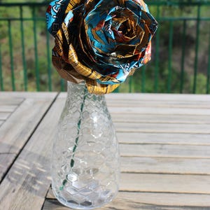 May include: A unique, handmade rose crafted from colorful, patterned materials, displayed in a clear glass vase with a honeycomb design. The rose is the focal point, with vibrant blues, oranges, and yellows. The vase sits on a wooden table.