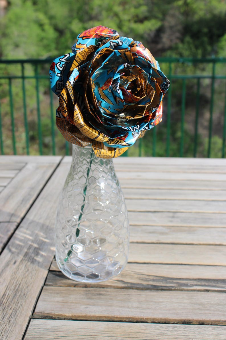 May include: A unique, handmade rose crafted from colorful, patterned materials, displayed in a clear glass vase with a honeycomb design. The rose is the focal point, with vibrant blues, oranges, and yellows. The vase sits on a wooden table.