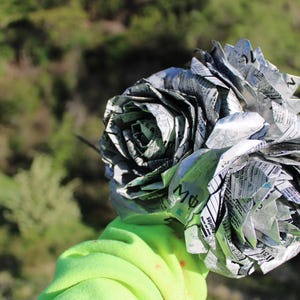 May include: A bouquet of paper roses made from newspaper. The roses are silver and gray, with visible text from the newspaper. The background is a blurred view of green trees and foliage.