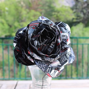 May include: A handcrafted rose made from recycled aluminum cans, primarily black with red and white accents. The flower is displayed in a clear glass vase, with a green background.