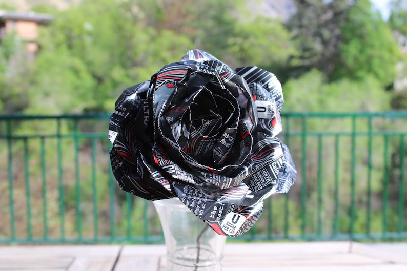 May include: A handcrafted rose made from recycled aluminum cans, primarily black with red and white accents. The flower is displayed in a clear glass vase, with a green background.