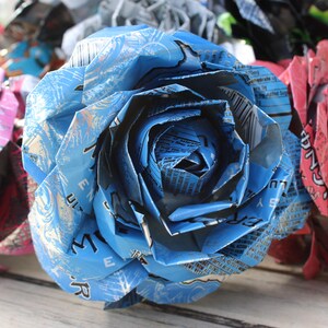 May include: Close-up of handmade paper flowers crafted from recycled aluminum cans. The central flower is blue, with others in pink and other colors. The petals are intricately folded, showcasing the can's original printed designs.