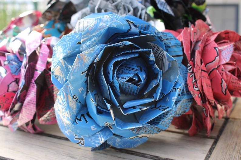 May include: Close-up of handmade paper flowers crafted from recycled aluminum cans. The central flower is blue, with others in pink and other colors. The petals are intricately folded, showcasing the can's original printed designs.