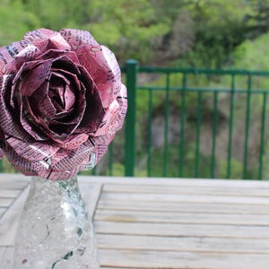 May include: A large, handcrafted rose made from pink and silver aluminum can material. The flower is displayed in a clear glass vase on a wooden table, with a green background of trees and a fence.