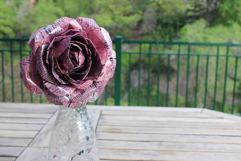 May include: A large, handcrafted rose made from pink and silver aluminum can material. The flower is displayed in a clear glass vase on a wooden table, with a green background of trees and a fence.