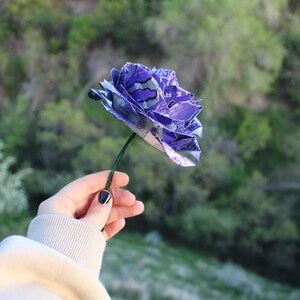 May include: A handmade purple flower crafted from recycled materials, held against a blurred green background. The flower's petals are made from a patterned material, and it has a green stem. The flower is a unique, eco-friendly decorative item.