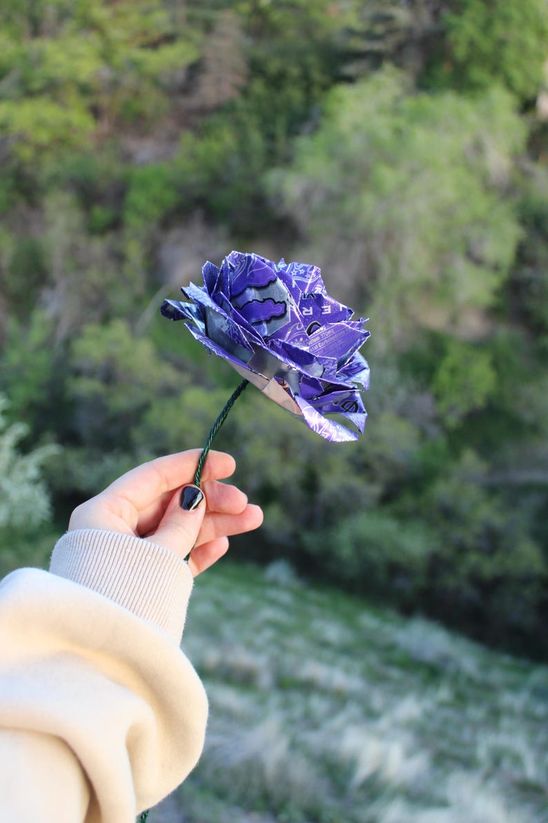 May include: A handmade purple flower crafted from recycled materials, held against a blurred green background. The flower's petals are made from a patterned material, and it has a green stem. The flower is a unique, eco-friendly decorative item.