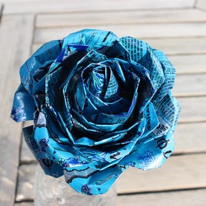 May include: A handcrafted, large blue rose made from recycled materials. The petals are intricately folded and layered, creating a realistic floral design. The rose is displayed in a clear glass vase, set against a wooden background.
