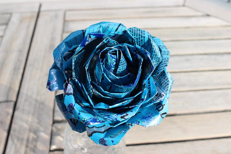 May include: A handcrafted, large blue rose made from recycled materials. The petals are intricately folded and layered, creating a realistic floral design. The rose is displayed in a clear glass vase, set against a wooden background.