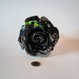 May include: A handcrafted black rose made from recycled aluminum cans. The rose has a detailed petal design with green accents and is displayed on a white surface. A quarter is next to the rose for size comparison.