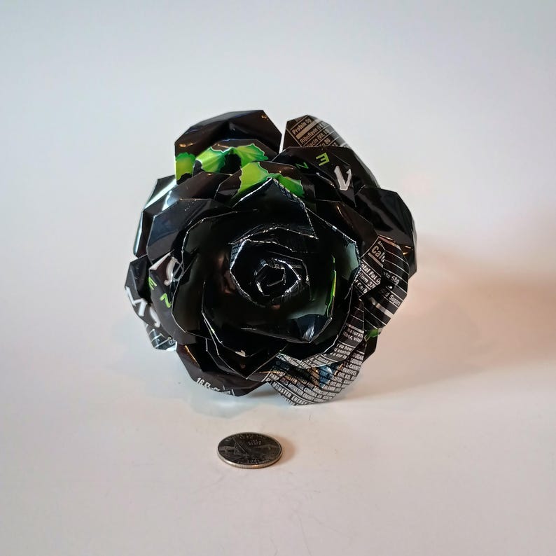 May include: A handcrafted black rose made from recycled aluminum cans. The rose has a detailed petal design with green accents and is displayed on a white surface. A quarter is next to the rose for size comparison.
