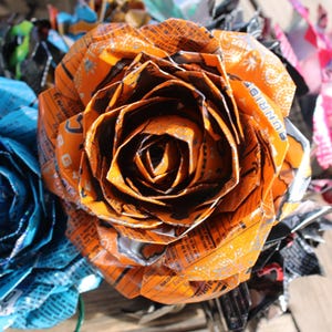 May include: Close-up of handmade paper roses in shades of orange, blue, and pink. The orange rose is in focus, showcasing intricate folds and textures. The flowers appear to be made from recycled materials.