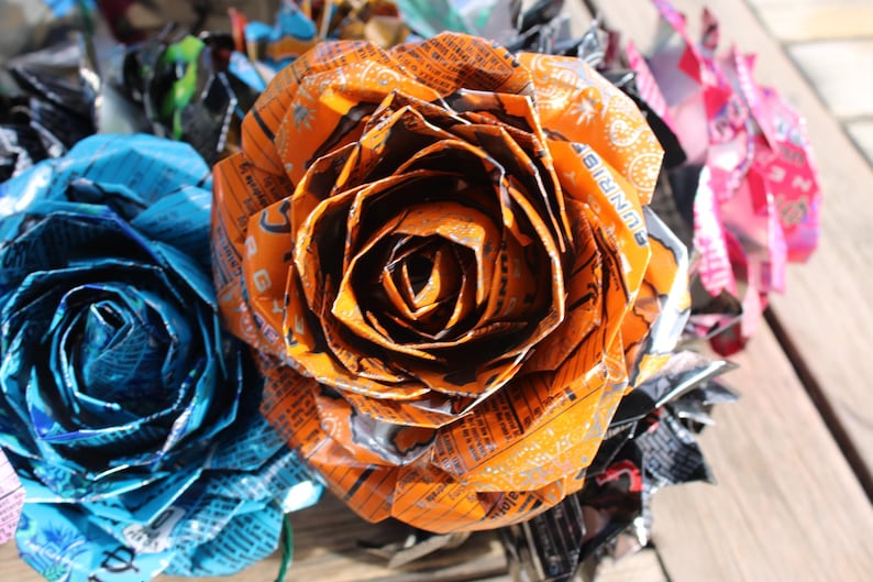 May include: Close-up of handmade paper roses in shades of orange, blue, and pink. The orange rose is in focus, showcasing intricate folds and textures. The flowers appear to be made from recycled materials.
