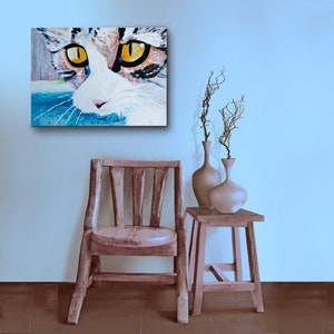 May include: A cat portrait painting with striking yellow eyes and a white and brown face hangs on a light blue wall. A wooden chair and small table with vases and branches complete the scene.