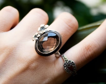 Könnte beinhalten: Ein silberner Ring mit einem großen ovalen Rauchquarzstein und drei kleineren blütenförmigen Akzenten. Der Ring wird an einem Finger getragen.