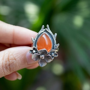 Könnte beinhalten: Ein silberner Ring mit einem tropfenförmigen orangefarbenen Edelstein, umgeben von floralen Details.