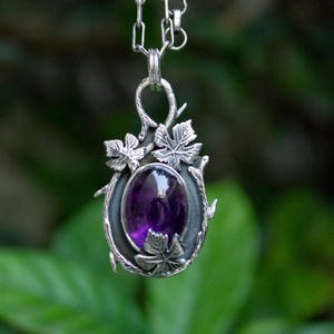 Può includere: Una collana con pendente in argento con una gemma ovale viola scuro. Il pendente è incorniciato da un metallo scuro e decorato con dettagli di foglie d'argento. La catena è a maglie delicate.