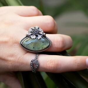 May include: A silver ring with a green gemstone and a flower design. The ring is on a finger.