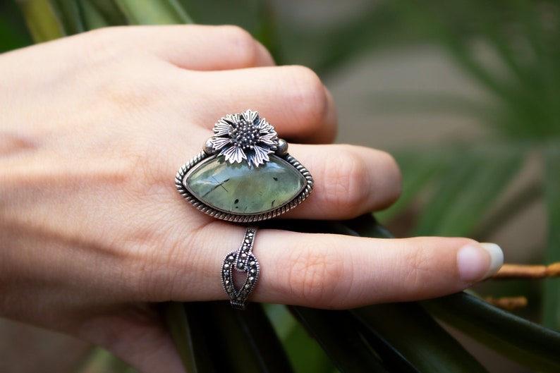 May include: A silver ring with a green gemstone and a flower design. The ring is on a finger.