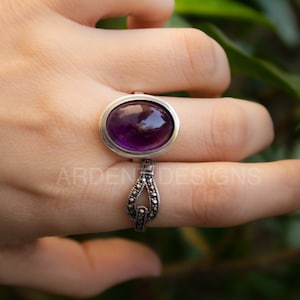 May include: A silver ring featuring a large oval amethyst gemstone set in a bezel setting. The band is adorned with a delicate marcasite design.
