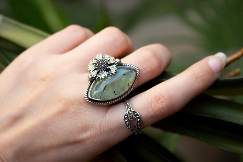 May include: A silver ring with a green gemstone and a silver flower design. The ring is on a finger.