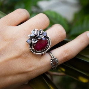 May include: A silver ring with a large red gemstone surrounded by smaller white gemstones and a silver flower design.
