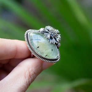 May include: A silver ring with a green gemstone and a flower design. The ring is set in a silver band with a rope-like design.