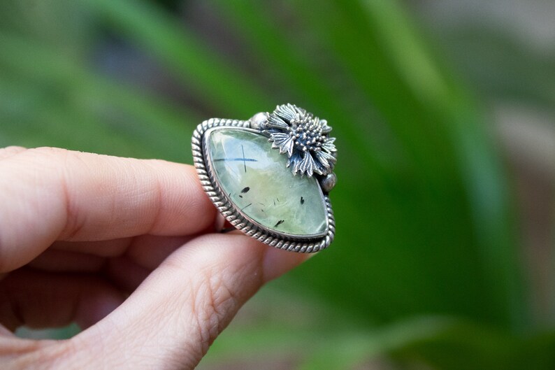 May include: A silver ring with a green gemstone and a flower design. The ring is set in a silver band with a rope-like design.