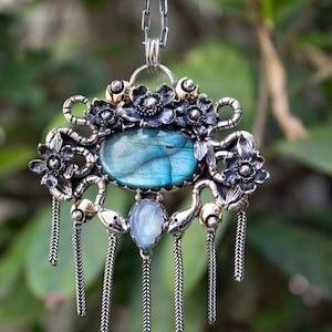 Puede incluir: Un colgante de plata con una gran piedra de labradorita ovalada de iridiscencia azul, una piedra de luna en forma de pera y detalles florales de metal. La pieza incluye cinco cadenas de plata colgantes y una cadena de eslabones.