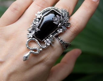 May include: A silver ring with a large, rectangular black stone. The ring has floral and leaf embellishments, and a coiled snake design. A distinctive piece of jewellery.