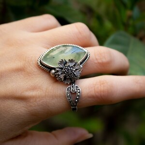 May include: A silver ring with a large green gemstone and a floral design. The ring is set on a finger.