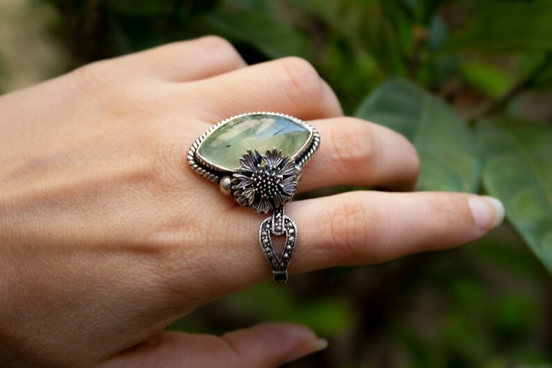 May include: A silver ring with a large green gemstone and a floral design. The ring is set on a finger.