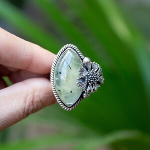 May include: A silver ring with a green gemstone and a silver flower design. The ring is set in a diamond shape.