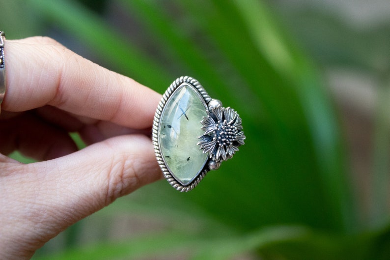 May include: A silver ring with a green gemstone and a silver flower design. The ring is set in a diamond shape.