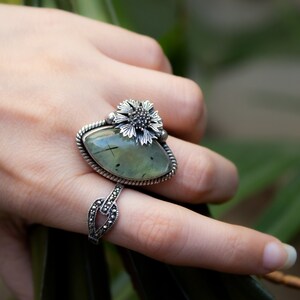 May include: A silver ring with a green gemstone and a flower design. The ring is on a finger.